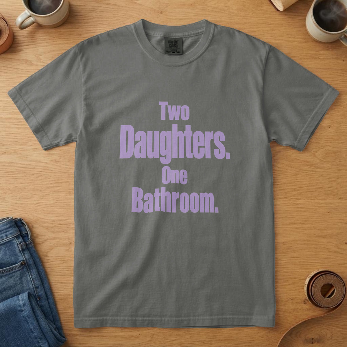 Pepper t-shirt with tall lavender type and the words "Two Daughters. One Bathroom.", on an oak table with a coffee mug