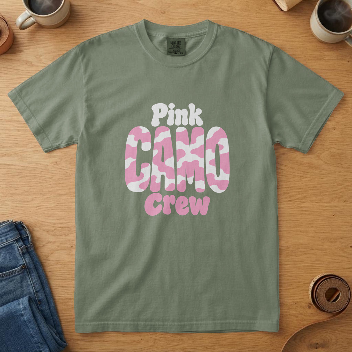 Moss t-shirt with pink and white camo-filled lettering and the words "Pink Camo Crew", on an oak table with a coffee mug