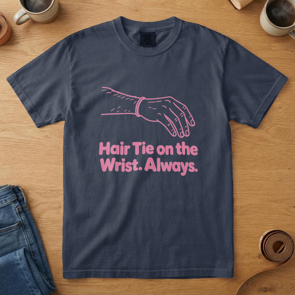 Navy t-shirt with a pink line drawing of a forearm with a hair tie and the words "Hair Tie on the Wrist. Always."