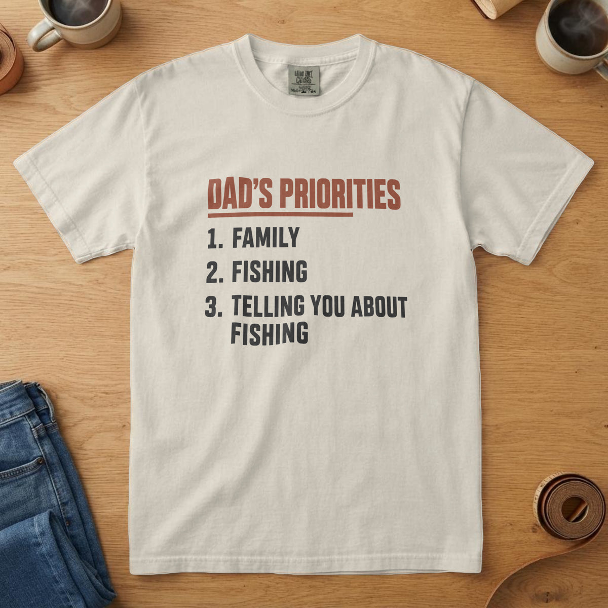 Ivory t-shirt with a list reading "Dad's Priorities: 1. Family 2. Fishing 3. Telling you about fishing", on an oak table