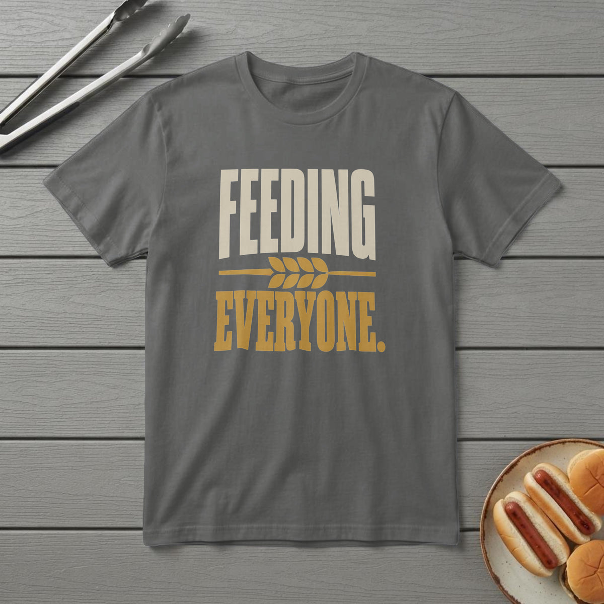 Pepper t-shirt with bold cream and mustard type reading "Feeding Everyone." and a wheat divider, on a grey deck
