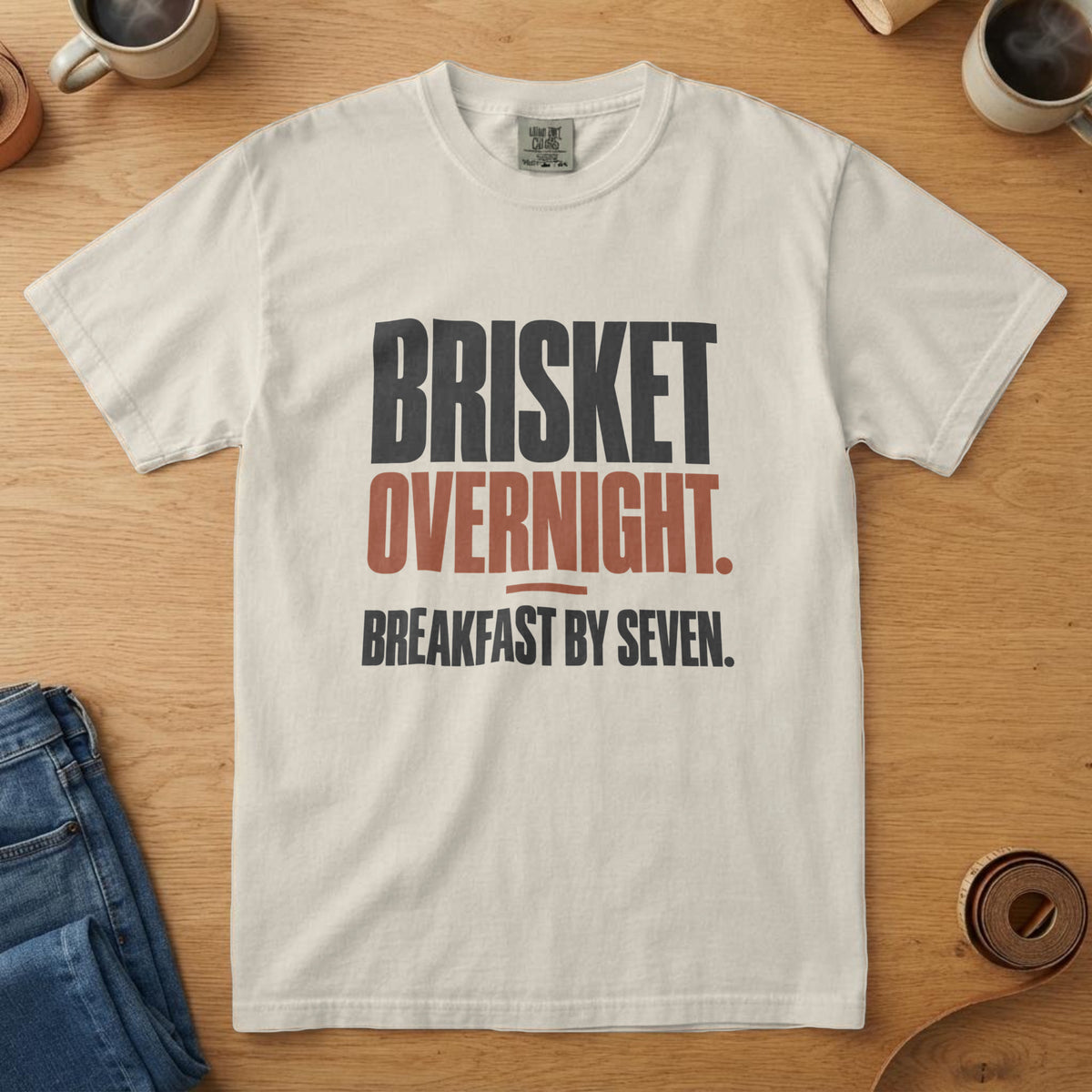 Ivory t-shirt with bold black and rust type reading "Brisket Overnight. Breakfast by Seven.", on an oak table