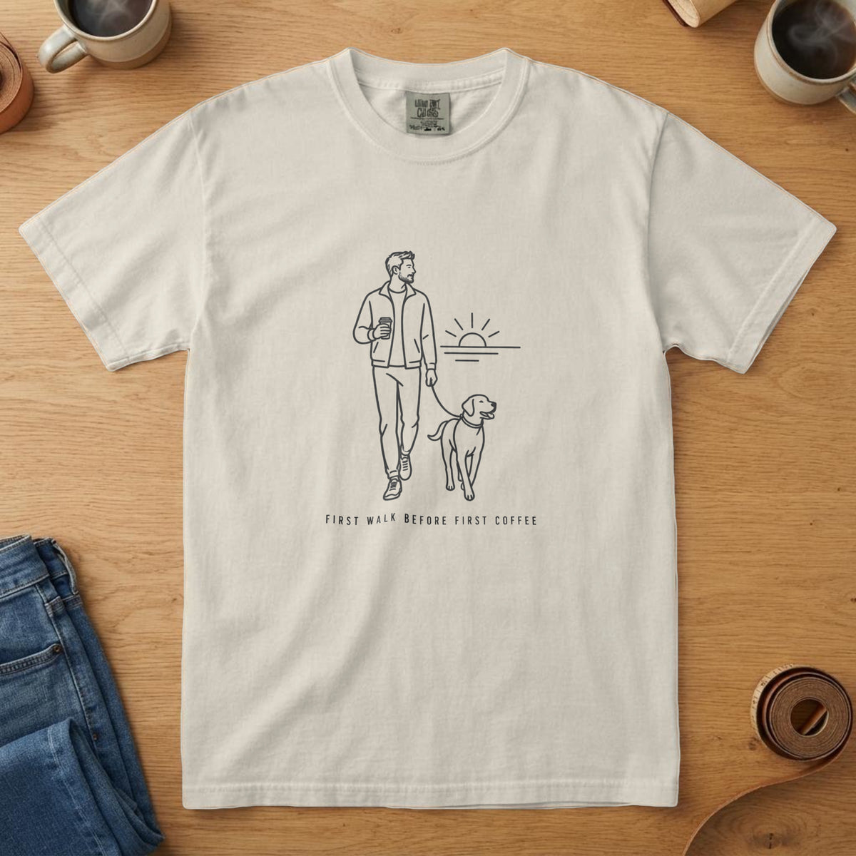 Ivory t-shirt with a near-black line drawing of a man walking a dog at dawn with a mug and "First Walk Before First Coffee"
