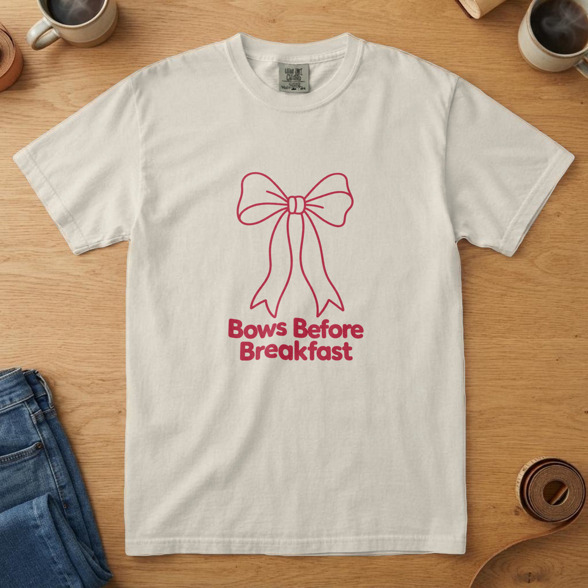 Ivory t-shirt with a red line drawing of a hair bow and the words "Bows Before Breakfast", on an oak table with a coffee mug