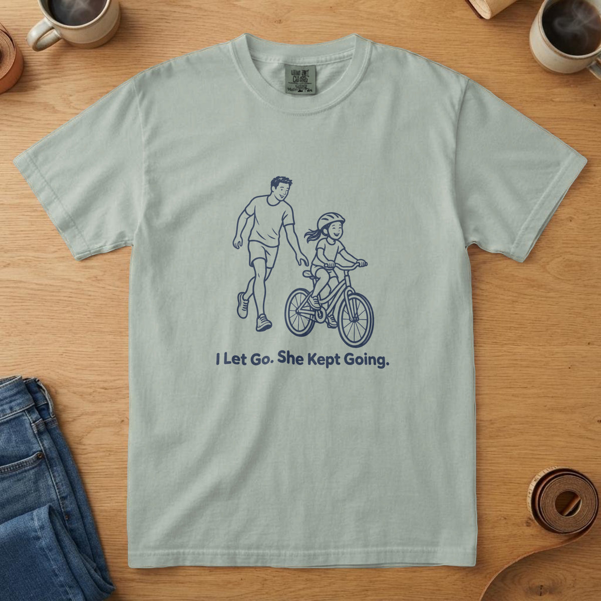 Bay t-shirt with a navy line drawing of a dad letting go of a girl's bike and the words "I Let Go. She Kept Going."
