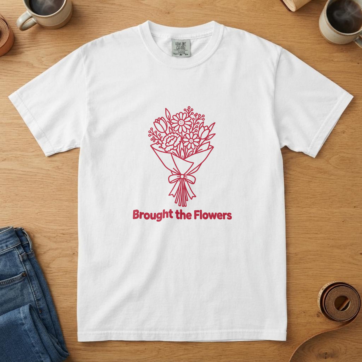 White t-shirt with a red line drawing of a bouquet and the words "Brought the Flowers", on an oak table with a coffee mug