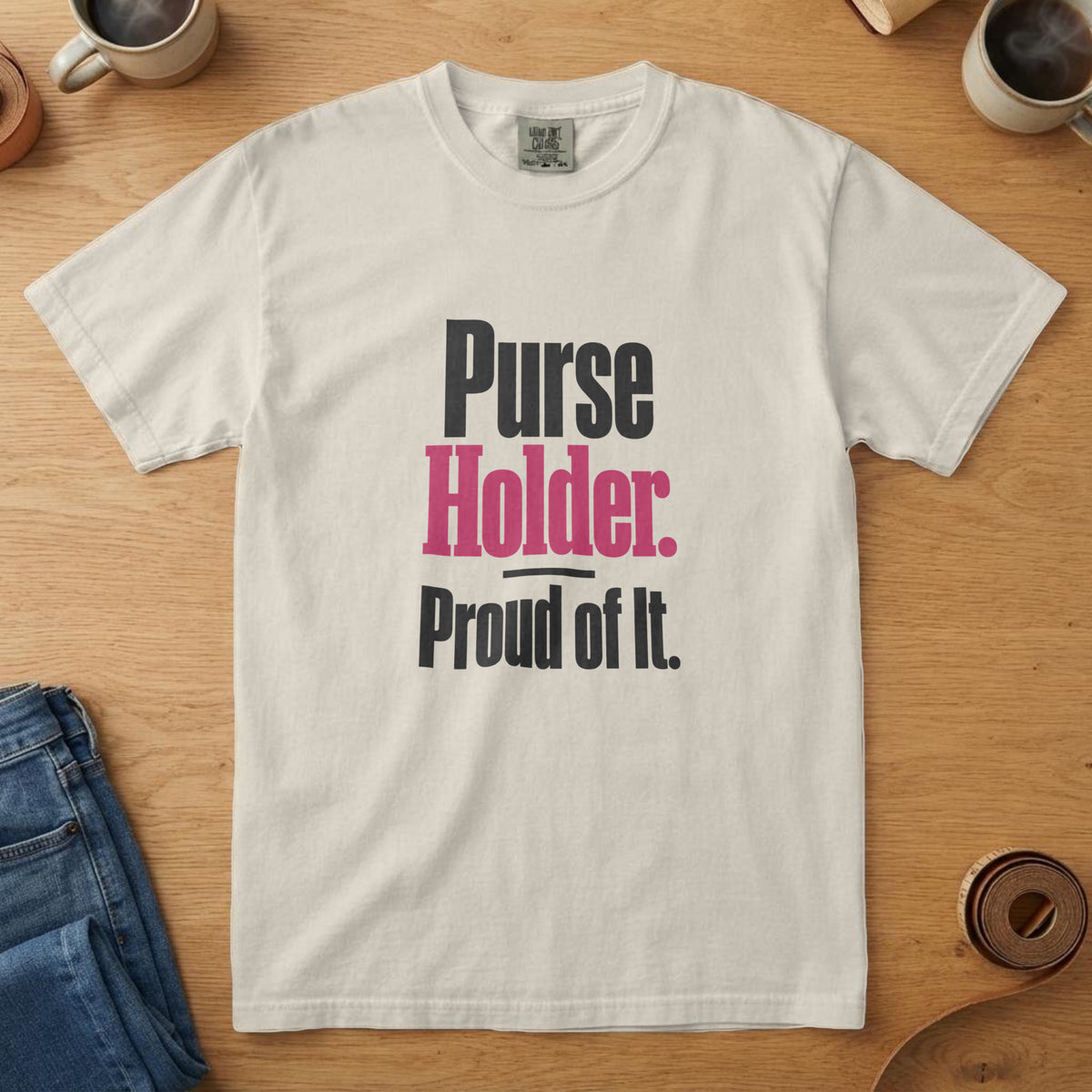 Ivory t-shirt with bold black and pink type and the words "Purse Holder. Proud of It.", on an oak table with a coffee mug