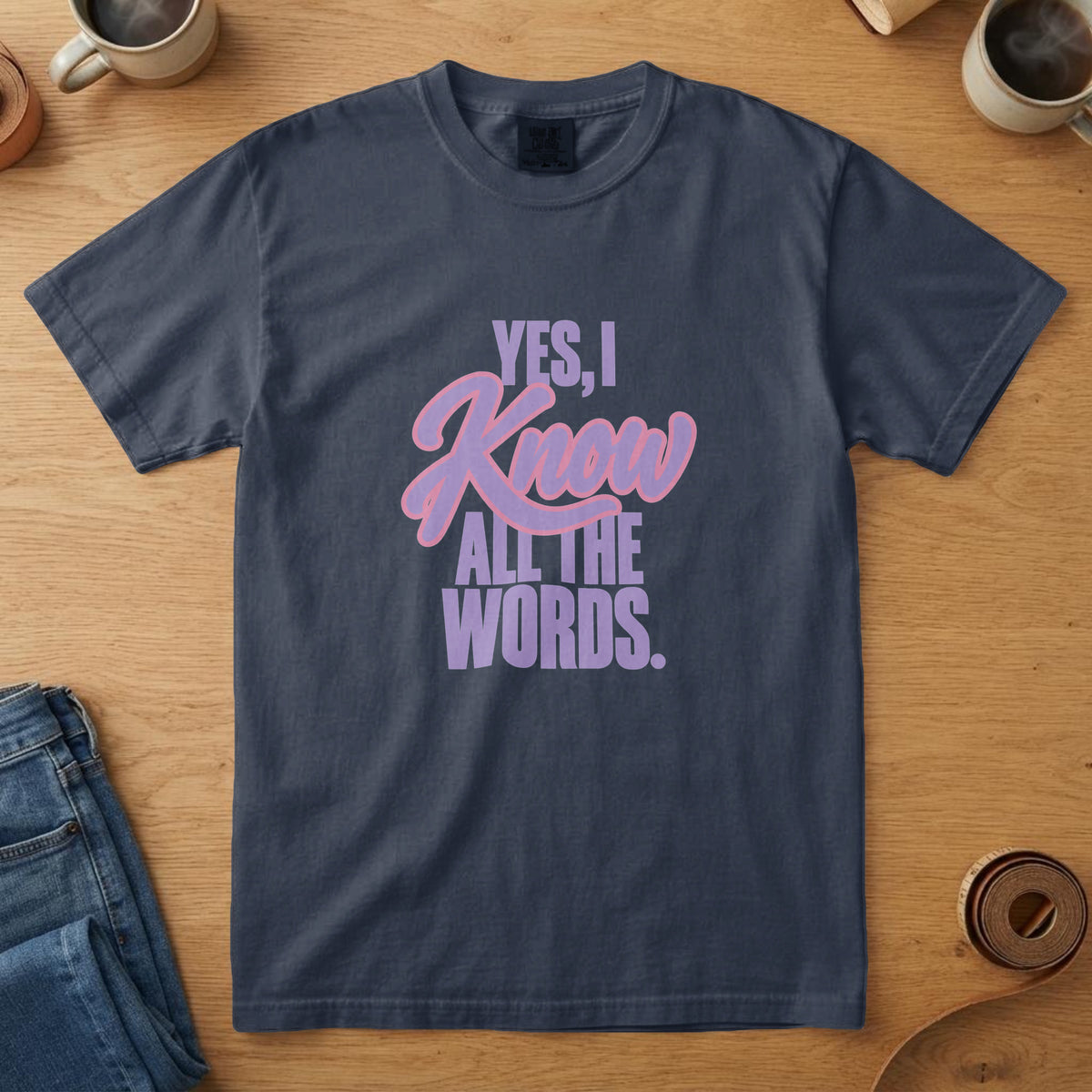 Navy t-shirt with lavender and pink lettering and the words "Yes, I Know All the Words.", on an oak table with a coffee mug