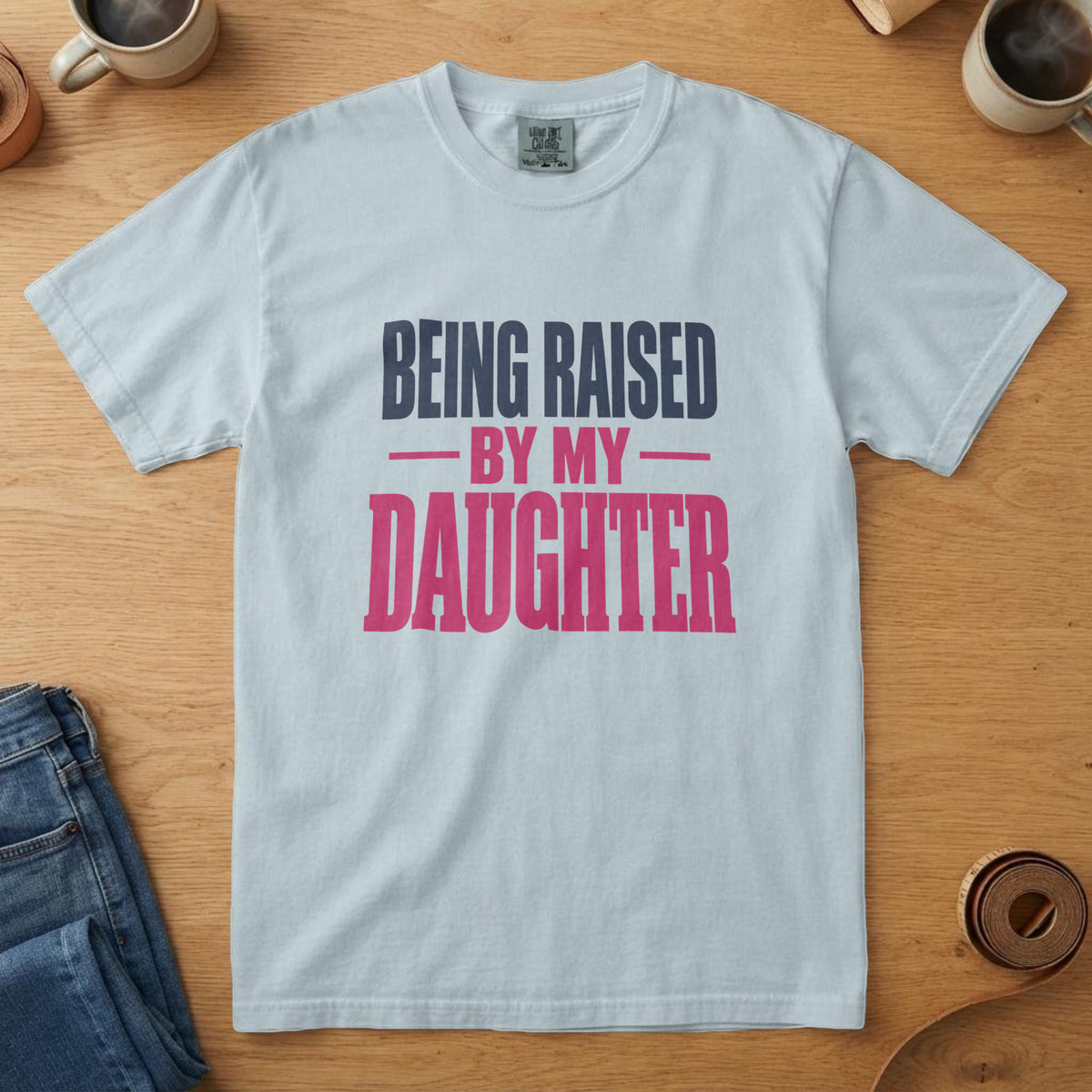 Chambray t-shirt with tall navy and pink type and the words "Being Raised by My Daughter", on an oak table with a coffee mug