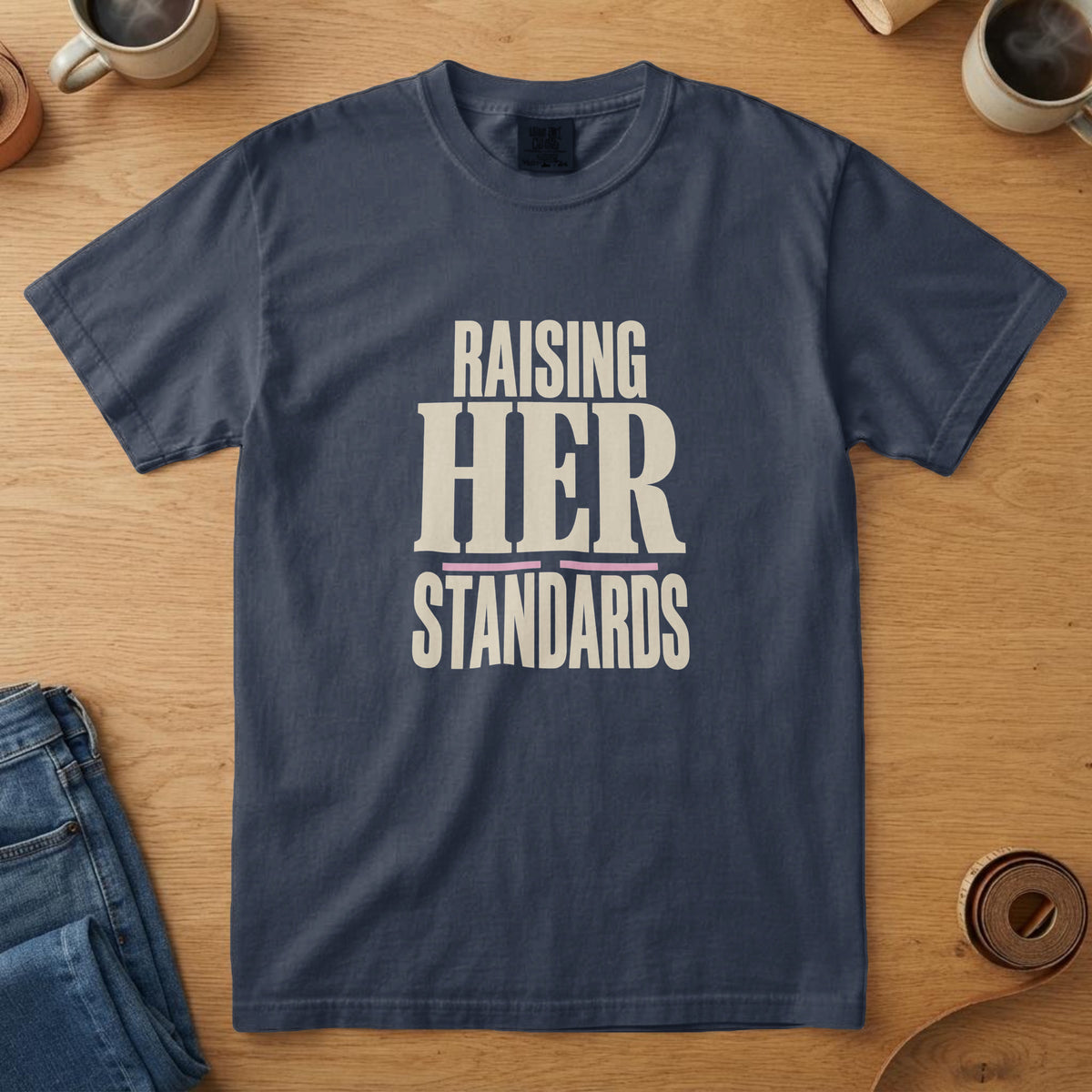 Navy t-shirt with bone serif type with a pink rule and the words "Raising Her Standards", on an oak table with a coffee mug