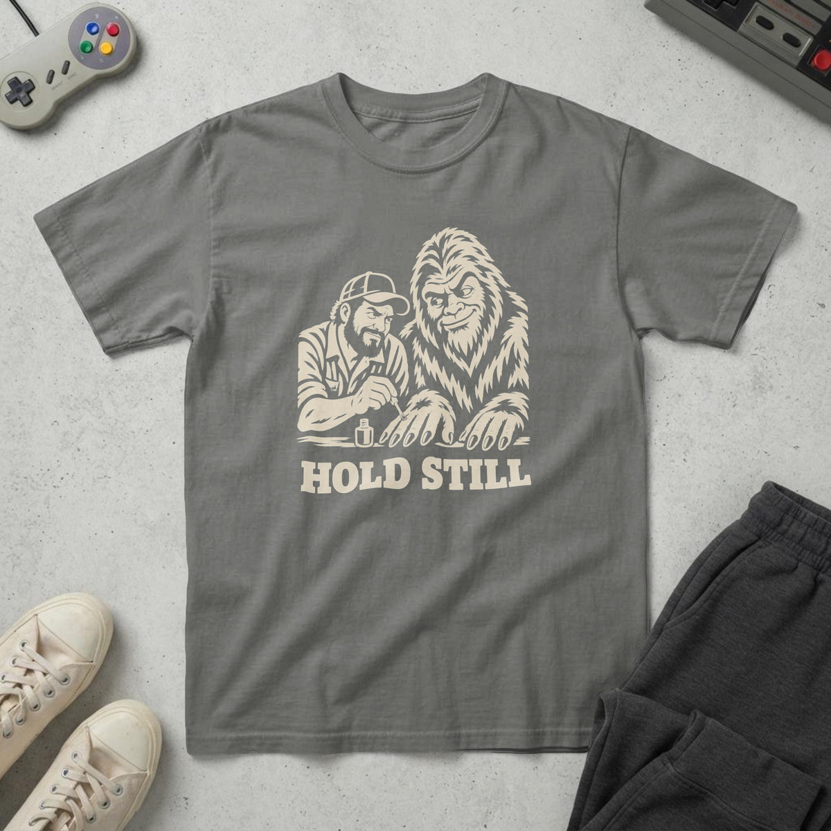 Pepper t-shirt with a cream woodcut of a dad painting a Sasquatch's claws and the words "Hold Still", on concrete
