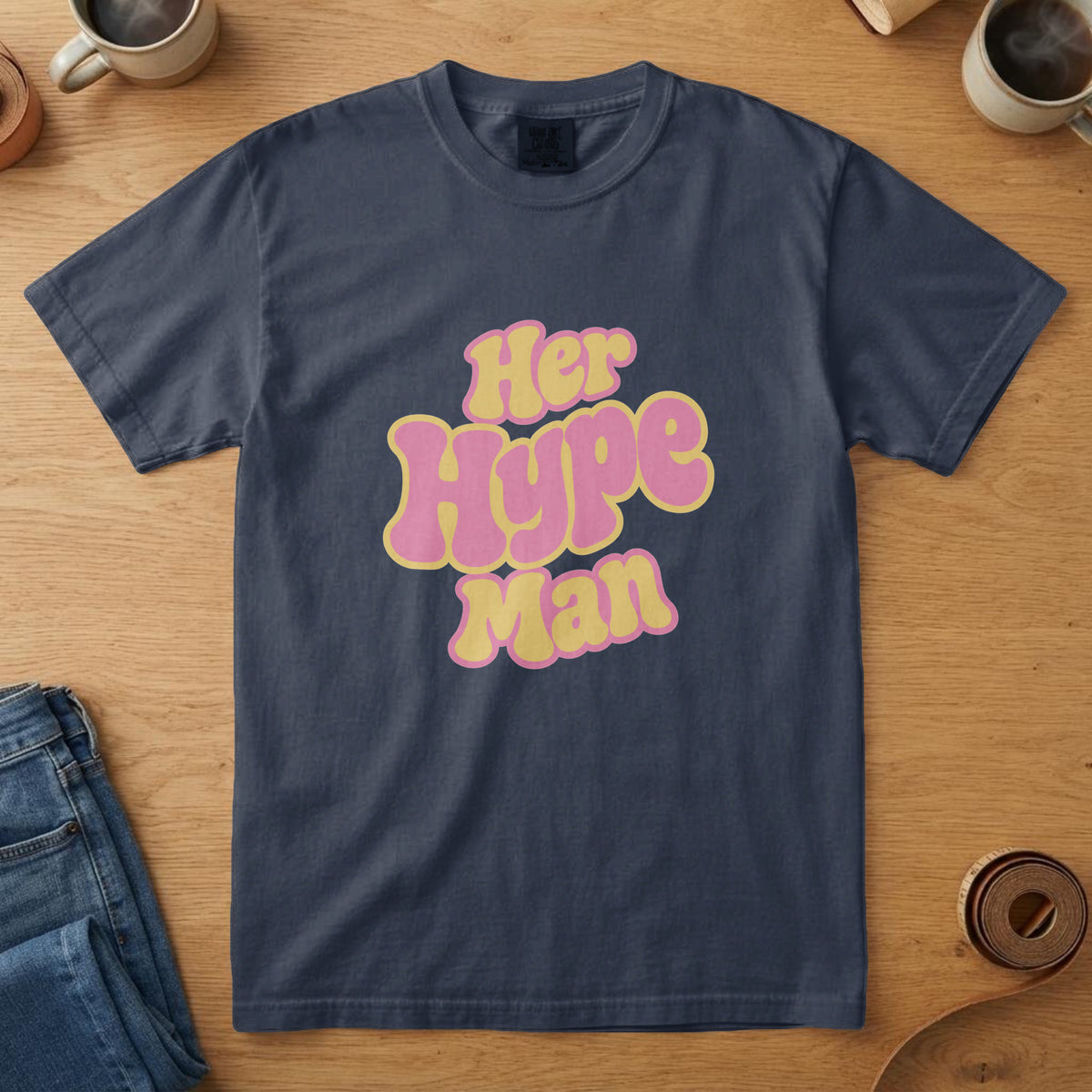 Navy t-shirt with chunky pink and yellow lettering and the words "Her Hype Man", on an oak table with a coffee mug